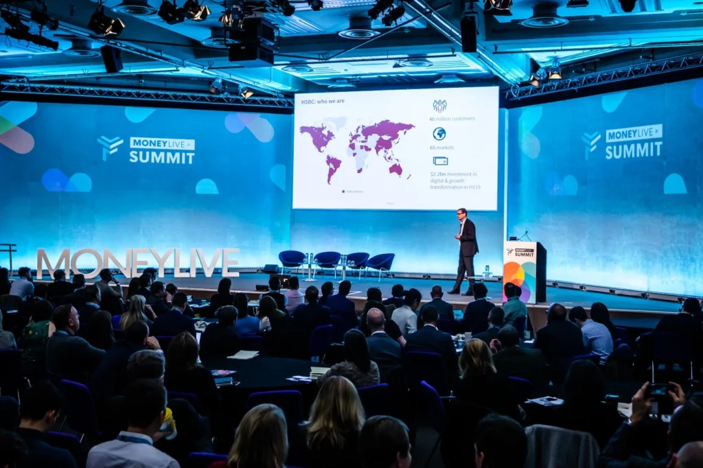 Speaker presenting on stage at MoneyLIVE Summit fintech conference 2023 with audience and large presentation screen discussing digital banking and payments.